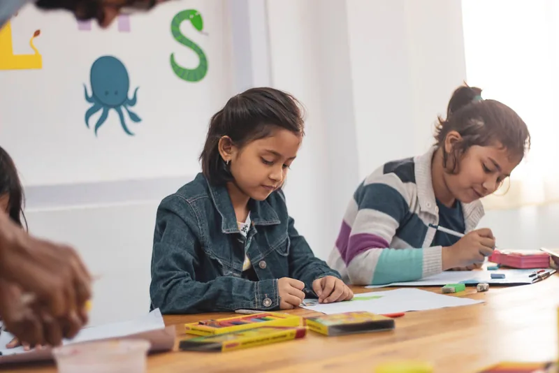 Two girls draw together at a table in a bright community classroom.