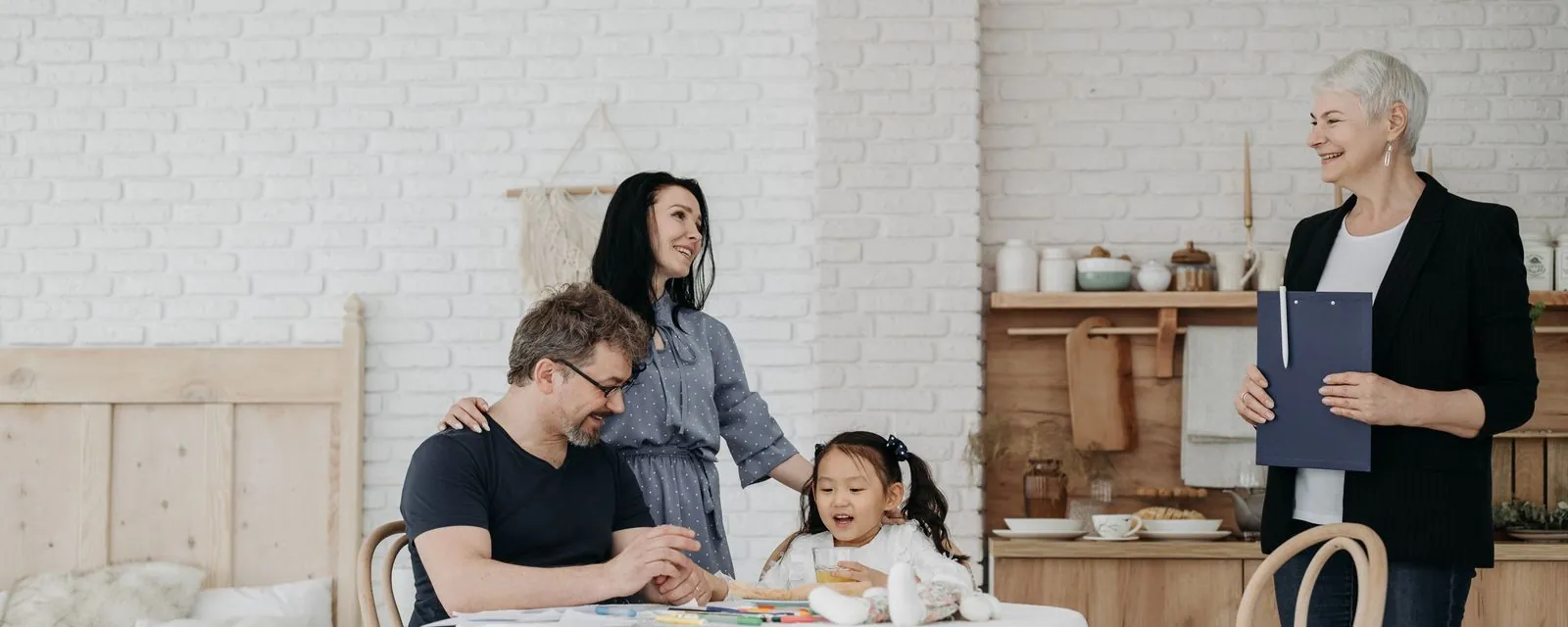 A staff member with a clipboard visits a family at their kitchen table while their daughter draws.