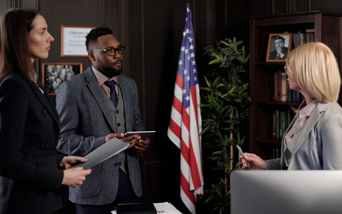 Three federal employees talk through a request in a wood-paneled office beside an American flag.