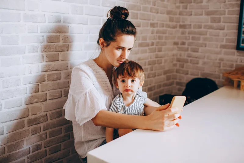 A mother holds her young son while looking at a phone at home.