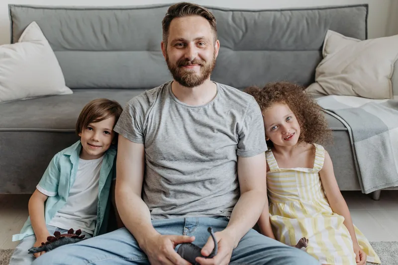 A father sits on the floor at home with his two children.